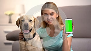 Happy woman hugging cute labrador dog, showing green screen smartphone, pet app