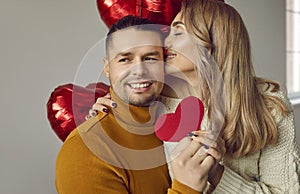 Happy woman hugs her man, kisses him and gives him a red heart shaped Valentine card