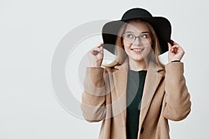 Happy woman with keen eyes and gentle smile wearing retro hat, eyeglasses and coat, holding sides of her hat. Flitry