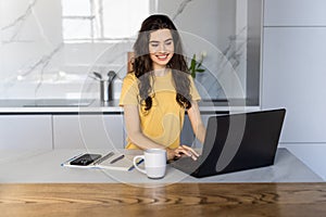 Happy young woman in the kitchen reading he news on her laptop