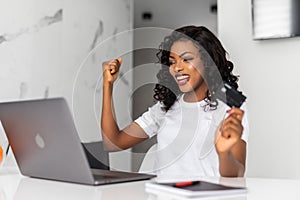 Happy woman with laptop with win gesture, credit cards and shopping bags at home