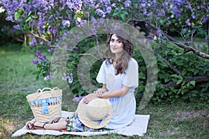 Happy Woman Life Style, portrait of a beautiful girl in a straw hat sitting near a blossoming lilac in the rays of the setting sun