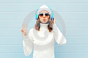 Happy woman listens to music in headphones wearing a sunglasses, knitted hat, sweater over blue background, blowing lips