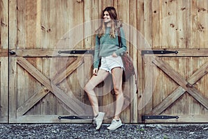 Happy woman with long legs look to the side near barn on the farm wearing casual outfit with shorts, backpack and sneakers.