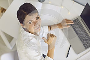 Happy woman looking at camera, smiling and showing thumbs-up while working on laptop