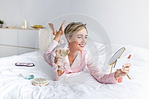 Happy woman lying in bed drinking alcohol taking selfie