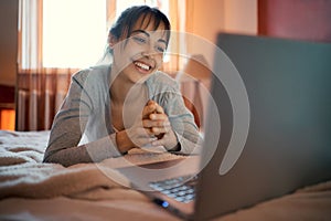 Happy woman lying on bed at home and chatting with boyfriend, having video call on laptop computer, using wireless speed
