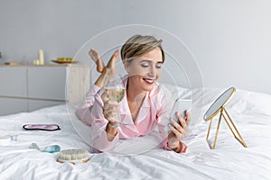 Happy woman lying in bed reading text message drinking alcohol