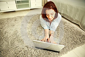 Happy woman lying on the carpet and typing on the laptop