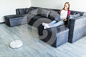 People, housework and technology concept. Happy woman lying on the couch while robot vacuum cleaner clean home