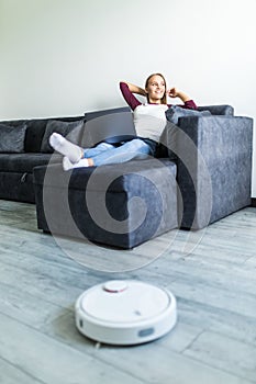 People, housework and technology concept. Happy woman lying on the couch while robot vacuum cleaner clean home