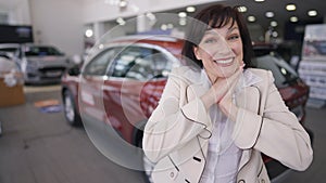 Happy woman making excited facial expression standing on the right with blurred red new car at background. Portrait of