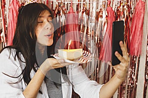 Happy woman making a wish and blowing candle on birthday cake and holding phone with video call