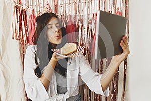 Happy woman making a wish and blowing candle on birthday cake and holding tablet with video call