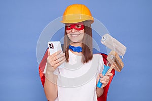 Happy woman painter wearing superhero costume and protective helmet holding painting roller and brush isolated over blue