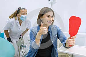 Happy woman patient looking in the mirror at the teeth, sitting in the dental chair