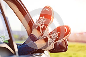 Happy woman put her legs out on open windowed car against blue sky and sun.