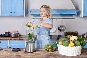 Happy woman putting ingredients into blender to make healthy smoothie for breakfast, cooking vegetables preparing salad