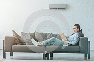 happy woman reading book on couch air conditioner on wall
