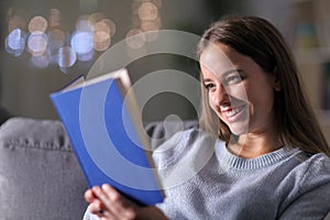 Happy woman reading a paper book on a couch in the night