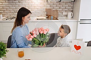 Happy woman receiving flowers and greeting card from her son at home. Mother\'s day celebration