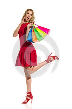 Happy Woman In Red Dress Is Standing On One Leg, Holding Colorful Shopping Bags And Shouting