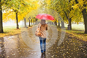 Happy woman with red umbrella walking at the rain in beautiful autumn park.