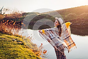 Happy woman relaxing by autumn lake at sunset. Stylish girl in hat plays with wind and scarf enjoying fall landscape