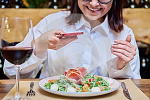 Happy woman in restaurant taking pictures of food with smartphone