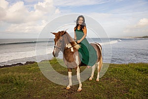 Happy woman riding horse near the ocean. Outdoor activities. Asia woman wearing long green dress. Traveling concept. Cloudy sky.