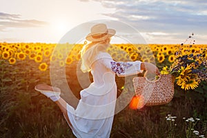 Happy woman running in blooming sunflower field at sunset having fun holding bag with flowers. Summer time in Ukraine