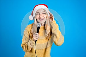 Happy woman in Santa Claus hat singing with microphone on blue background. Christmas music