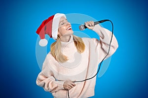 Happy woman in Santa Claus hat singing with microphone on blue background. Christmas music