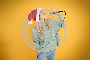 Happy woman in Santa Claus hat singing with microphone on yellow background. Christmas music