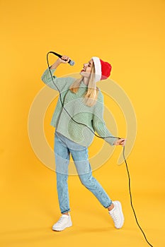 Happy woman in Santa Claus hat singing with microphone on yellow background. Christmas music