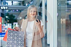 Happy woman shopper in the supermarket uses the phone, senior pensioner holds in her hands colored shopping bags