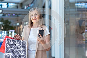 Happy woman shopper in the supermarket uses the phone, senior pensioner holds in her hands colored shopping bags