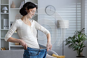 Happy woman showing weight loss by wearing old jeans at home