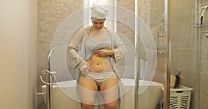 Happy woman sit on bath in bathrobe and head towel massaging clean body with brush. Anticellulite dry massage of belly.