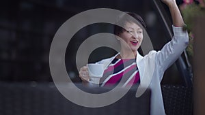 Happy woman sits in a cafe with a cup of coffee and greets her friends on the street