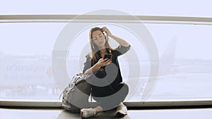 Happy woman sits with smartphone by airport window. Caucasian girl with backpack using messenger app in terminal. 4K.