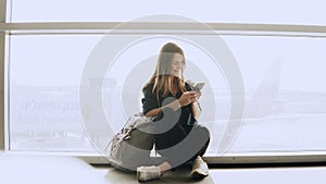 Happy woman sits with smartphone by airport window. Caucasian girl with backpack using messenger app in terminal. 4K.