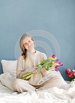 Happy woman sitting on the bed wearing pajamas holding tulip flowers bouquet, blue wall background