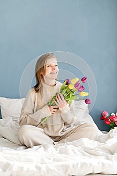 Happy woman sitting on the bed wearing pajamas holding tulip flowers bouquet, blue wall background