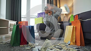 Happy woman is sitting on a carpet in a cozy room among shopping bags and making money rain from US dollar bills