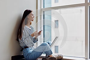 Happy woman sitting on the floor by the windows with a laptop on her lap and holding her phone, a view of the big city