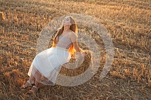 Happy woman sitting on hay stack walking in summer evening, beautiful romantic girl with long hair outdoor in field at sunset