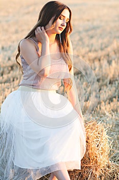 Happy woman sitting on hay stack walking in summer evening, beautiful romantic girl with long hair outdoors in field at sunset