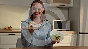 Happy woman sitting at the kitchen table chooses between a cake and a vegetable salad, chooses a cake