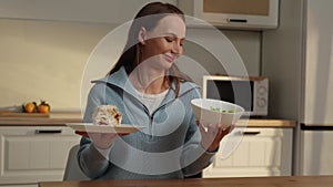 Happy woman sitting at the kitchen table chooses between a cake and a vegetable salad, chooses a salad